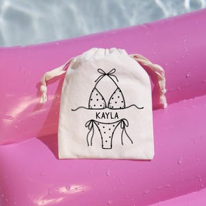 May include: A small off-white cotton drawstring pouch featuring a black line art graphic of a polka-dot bikini and the name KAYLA printed in the center. The bag has two cream-colored fabric drawstrings at the top for closure and storage.