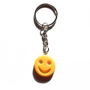 May include: A yellow smiley face keychain with a silver chain and ring. The smiley face has two eyes and a curved mouth, and is made of a smooth, solid material. The keychain is on a white background.