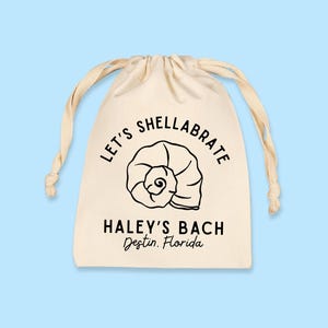 May include: A cream-colored drawstring bag with black text and a seashell graphic. The text reads "LET'S SHELLABRATE" above a seashell illustration, and "HALEY'S BACH Destin, Florida" below. The background is light blue.