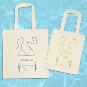 May include: Two canvas tote bags with white handles. One bag has a blue bikini outline with the name "BROOKLYN" below it. The other bag has a green bikini outline with the name "JEANA" below it. The bags are on a blue water background.