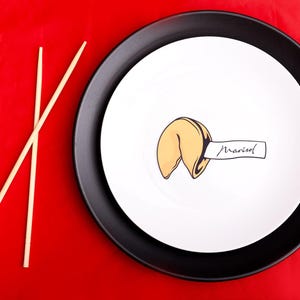 May include: A fortune cookie with the name "Marisol" on a white plate, set on a black plate, with chopsticks on a red background. The fortune cookie is light brown with a white paper strip.