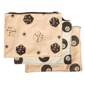 May include: Three rectangular zipper pouches with different patterns. One pouch has a beige background with dice and starburst designs. Another has a white background with black eight balls. The third pouch has a beige background with black eight balls.