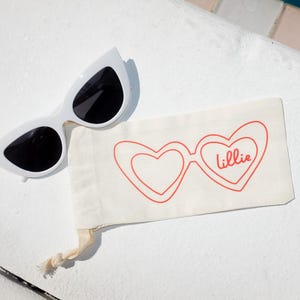 May include: White cat-eye sunglasses with dark lenses paired with a cream-colored fabric pouch. The pouch features a red line-art graphic of heart-shaped glasses with the name lillie written in cursive script inside one lens.