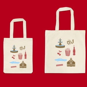 May include: Two off-white tote bags with cream-colored handles. Each bag features a collection of colorful illustrations, including a fountain, film reel, popcorn, cherries, a soda bottle, a car, a church, and a comb.