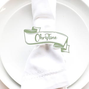 May include: A place setting with a white plate, napkin, fork, and knife. A green banner with the name "Christine" is wrapped around the napkin. The setting is on a white surface.