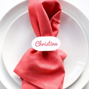 May include: A place setting with a folded coral napkin, a white oval name tag that reads "Christina", a fork, a knife, and two white plates. The setting is on a white surface.