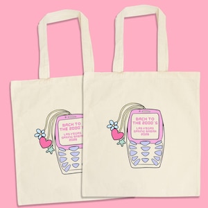 May include: Two off-white tote bags with a pink background. Each bag features a retro cell phone graphic with the text "Back to the 2000's Las Vegas Spring Break 2028". The phone is decorated with hearts and flowers.