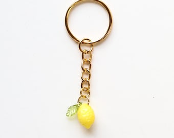 May include: A gold-colored keychain with a small, yellow lemon charm and a green leaf. The keychain has a gold ring and chain, and the lemon charm is detailed with a textured surface. The background is plain white.