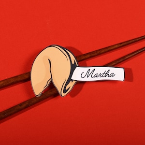 May include: A pair of dark brown wooden chopsticks positioned diagonally behind a paper cutout of a tan fortune cookie. A white paper strip extends from the fortune cookie with the name Martha printed in black cursive script on the white surface.