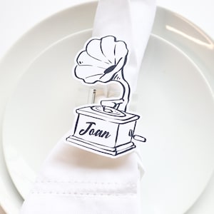 May include: A black and white place card featuring a vintage gramophone design with the name "Joan" printed on it. The place card is attached to a white napkin on a white plate, with a fork and knife visible.