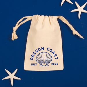 May include: A cream-colored drawstring bag with the text "OREGON COAST JULY 2026" in blue, along with a seashell graphic. Four white starfish are scattered around the bag on a blue background.