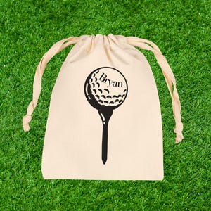 May include: A beige drawstring bag with a black golf ball and tee graphic. The name "Bryan" is printed above the golf ball. The bag is set against a green grass background.