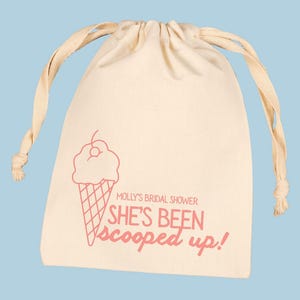 May include: A cream-colored drawstring bag with a pink ice cream cone graphic and the text "Molly's Bridal Shower She's Been Scooped Up!" on a light blue background. The bag has a rounded shape and a drawstring closure.