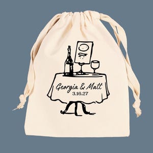 May include: A cream-colored drawstring bag with a black illustration of a table set for a meal. The table has a bottle, a menu, and a wine glass. The text "Georgia & Matt 3.16.27" is printed on the table.