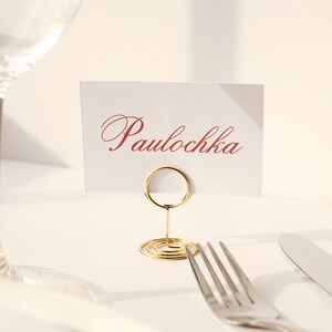 May include: A place setting with a wine glass, a card holder, a fork, and a knife. The card displays the name "Paulochka" in red script. The card holder is gold-colored and the utensils are silver.