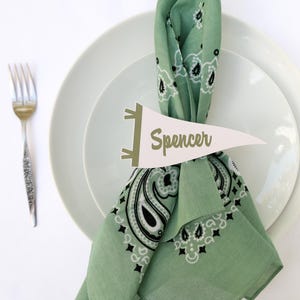 May include: A place setting with a green bandana-style napkin, a white pennant with the name "Spencer", and a silver fork and knife. The napkin is folded and placed on a white plate.