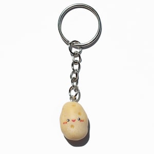 May include: A keychain featuring a small, light-colored potato charm with a smiling face and rosy cheeks. The charm is attached to a silver chain and keyring, set against a white background.