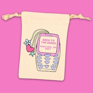 May include: A cream-colored drawstring bag featuring a cartoon flip phone design with the text "Back to the 2000s Portland, OR 2021". The phone is pink with blue buttons, and the bag has a pink background.