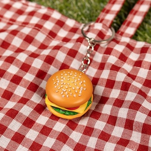May include: A red and white checkered tote bag with a burger keychain. The bag is lying on green grass.
