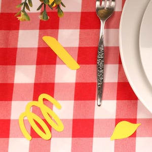 Puede incluir: Una mesa puesta sobre un mantel a cuadros rojos y blancos. El ajuste incluye platos blancos, una servilleta blanca doblada, un tenedor, un cuchillo y una copa de vino. Una tarjeta con el nombre "Anthony" está sobre la servilleta. Formas decorativas de pasta están dispersas.