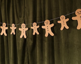 Puede incluir: Una guirnalda de figuras de hombre de jengibre con caras sonrientes y detalles de botones, ensartadas en una cuerda blanca. La decoración festiva se muestra contra una cortina verde oscuro, ideal para las fiestas.