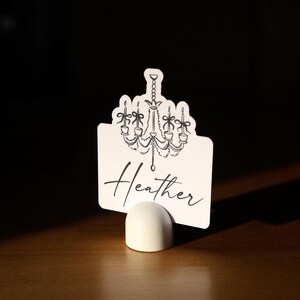 May include: A white place card with a black chandelier design and the name "Heather" in script. The card is held in a small, white, rounded holder, set against a dark background.