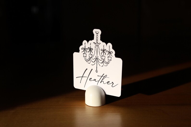 May include: A white place card with a black chandelier design and the name "Heather" in script. The card is held in a small, white, rounded holder, set against a dark background.