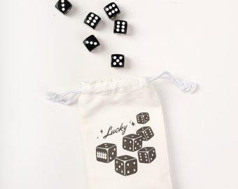 Puede incluir: Una bolsa blanca de cordón con la palabra "Lucky" impresa en ella. La bolsa está llena de dados negros con puntos blancos.
