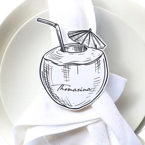 May include: A black and white coconut-shaped place card with a straw and umbrella, resting on a white napkin and plate. The name "Thomasina" is written on the coconut.