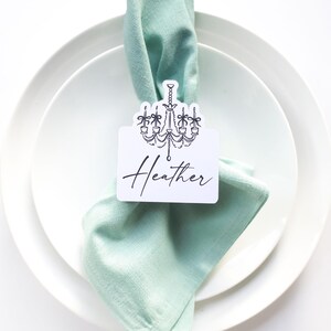 May include: A place setting with a white plate, a light green napkin, and a name card that says "Heather" with a chandelier design. A fork and knife are on either side of the plate.
