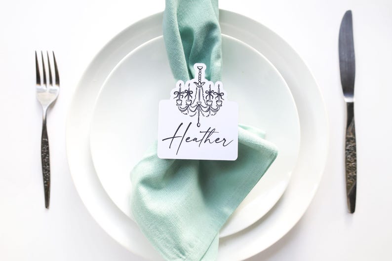May include: A place setting with a white plate, a light green napkin, and a name card that says "Heather" with a chandelier design. A fork and knife are on either side of the plate.