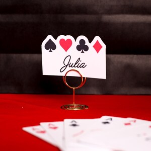 Puede incluir: Una tarjeta blanca con el nombre "Julia" y s&iacute;mbolos de naipes, sujeta por un soporte de alambre dorado. Los s&iacute;mbolos son picas, corazones, tr&eacute;boles y diamantes. Varias cartas de juego son visibles sobre una superficie roja.