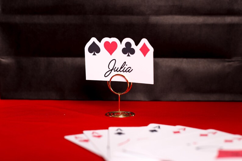 Puede incluir: Una tarjeta blanca con el nombre "Julia" y s&iacute;mbolos de naipes, sujeta por un soporte de alambre dorado. Los s&iacute;mbolos son picas, corazones, tr&eacute;boles y diamantes. Varias cartas de juego son visibles sobre una superficie roja.