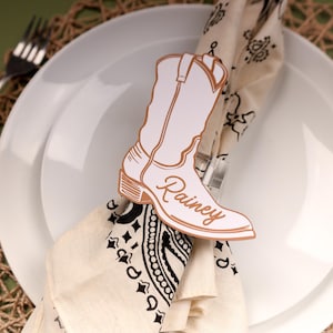 May include: A white plate with a folded napkin and a decorative cowboy boot place card. The place card is white with a brown outline and the name "Rainey" in script. A fork and knife are on either side of the plate.
