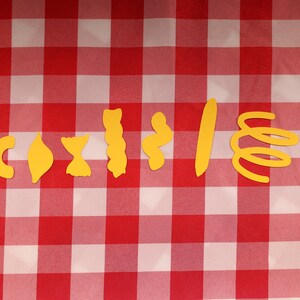 May include: A collection of yellow pasta shapes, including elbow macaroni, shells, bow ties, and spirals, arranged on a red and white checkered tablecloth. A U.S. quarter is included for size comparison.