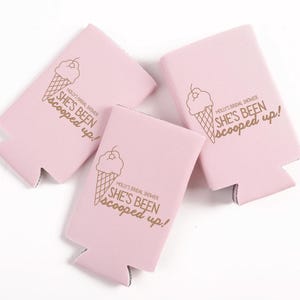 May include: Three pink can coolers with a gold ice cream cone graphic and the text "SHE'S BEEN scooped up!" printed on them. The text "MOLLY'S BRIDAL SHOWER" is also printed on the coolers. The coolers are made of a soft material.