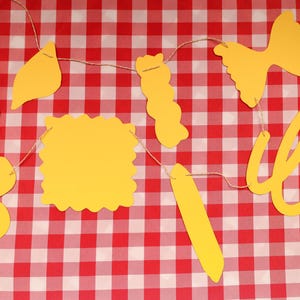 May include: A yellow pasta-themed banner with various pasta shapes, including farfalle, rotini, and lasagna, strung on twine. The banner is set against a red and white checkered tablecloth, creating a playful and thematic display.