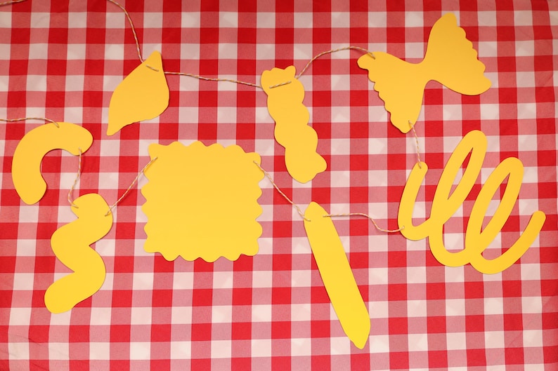 May include: A yellow pasta-themed banner with various pasta shapes, including farfalle, rotini, and lasagna, strung on twine. The banner is set against a red and white checkered tablecloth, creating a playful and thematic display.