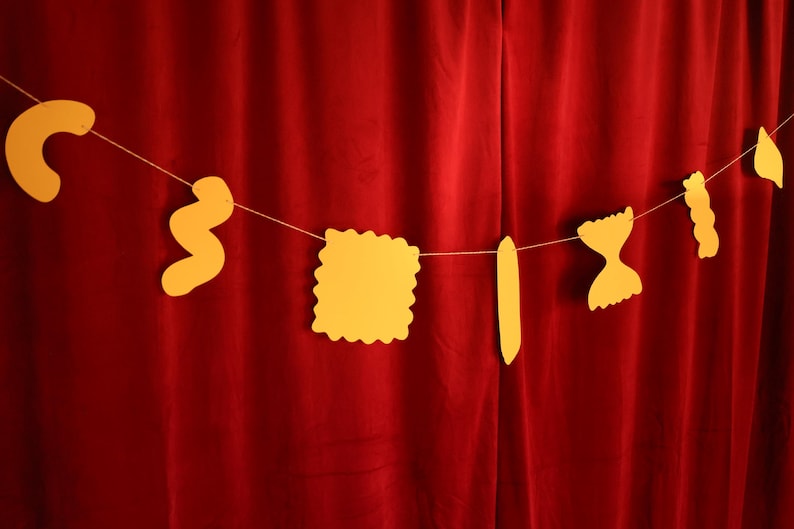 May include: A string of yellow pasta-shaped cutouts hangs against a red curtain backdrop. The pasta shapes include a curved macaroni, a wavy shape, a square, a long rectangle, a bow tie, a spiral, and a teardrop shape.