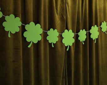 Puede incluir: Una guirnalda de recortes de tréboles verdes de papel ensartados en una cuerda blanca. Los tréboles se exhiben contra una cortina verde oscuro, creando una decoración festiva para el Día de San Patricio.