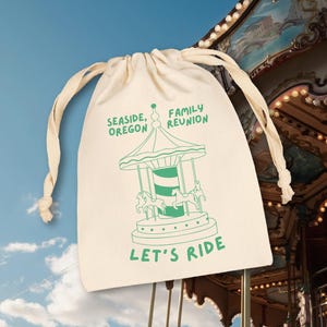 May include: A cream-colored fabric drawstring bag featuring a green graphic of a carousel. The text reads SEASIDE, OREGON FAMILY REUNION and LET'S RIDE. The bag has two off-white fabric cords at the top for closure and a simple rectangular shape.