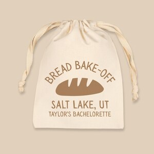 May include: A cream-colored drawstring bag with the words "Bread Bake-Off" and a bread loaf graphic printed in brown. The text "Salt Lake, UT" and "Taylor's Bachelorette" are also printed on the bag.