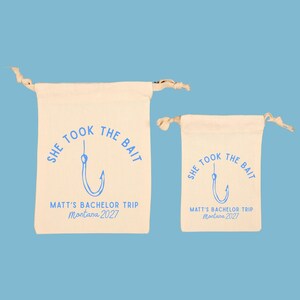May include: Two off-white cotton drawstring bags of different sizes, each featuring a blue graphic of a fishing hook and the text "SHE TOOK THE BAIT", "MATT'S BACHELOR TRIP", and "Montana 2027" printed in a blue sans-serif and script font.