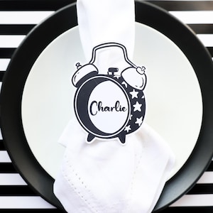 May include: A place setting with a white plate, black charger, and a napkin with a black and white alarm clock-shaped name tag that says "Charlie". A fork and knife are on either side of the plate. The background is a black and white striped pattern.