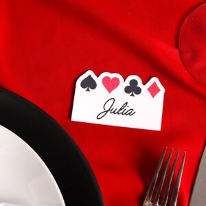 Puede incluir: Una tarjeta de sitio blanca con s&iacute;mbolos de naipes y el nombre "Julia" en escritura cursiva. La tarjeta est&aacute; sobre un mantel rojo, cerca de un plato, cubiertos y una copa de vino.