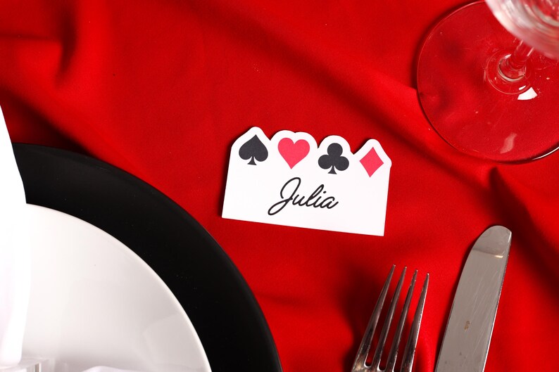 Puede incluir: Una tarjeta de sitio blanca con s&iacute;mbolos de naipes y el nombre "Julia" en escritura cursiva. La tarjeta est&aacute; sobre un mantel rojo, cerca de un plato, cubiertos y una copa de vino.