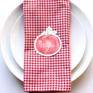 May include: A place setting with a red and white checkered napkin, a white plate, a fork, and a knife. A decorative tomato-shaped name tag with the name "Angie" is on the napkin. The setting is on a white surface.