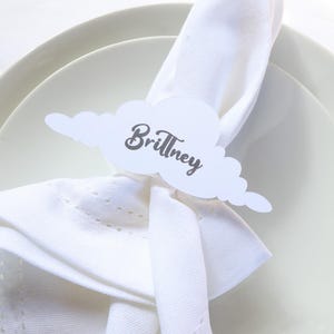 May include: A white cloud-shaped place card with the name "Brittney" in gray script sits atop a folded white napkin. The napkin is placed on a white plate, with another plate visible in the background. The scene suggests a wedding or event setting.