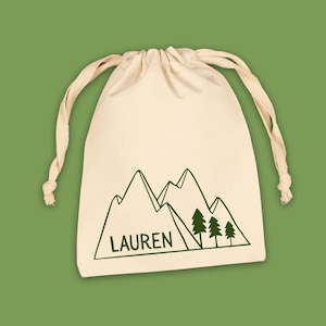 May include: A cream-colored drawstring bag with a green mountain and tree graphic. The name "LAUREN" is printed below the mountains. The bag is on a green background.