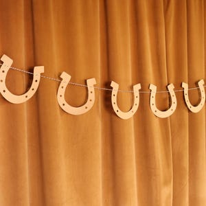 May include: A garland of light brown horseshoe cutouts strung on a thin white cord. The horseshoes are evenly spaced and hang against a backdrop of a brown curtain. The garland is a decorative item.